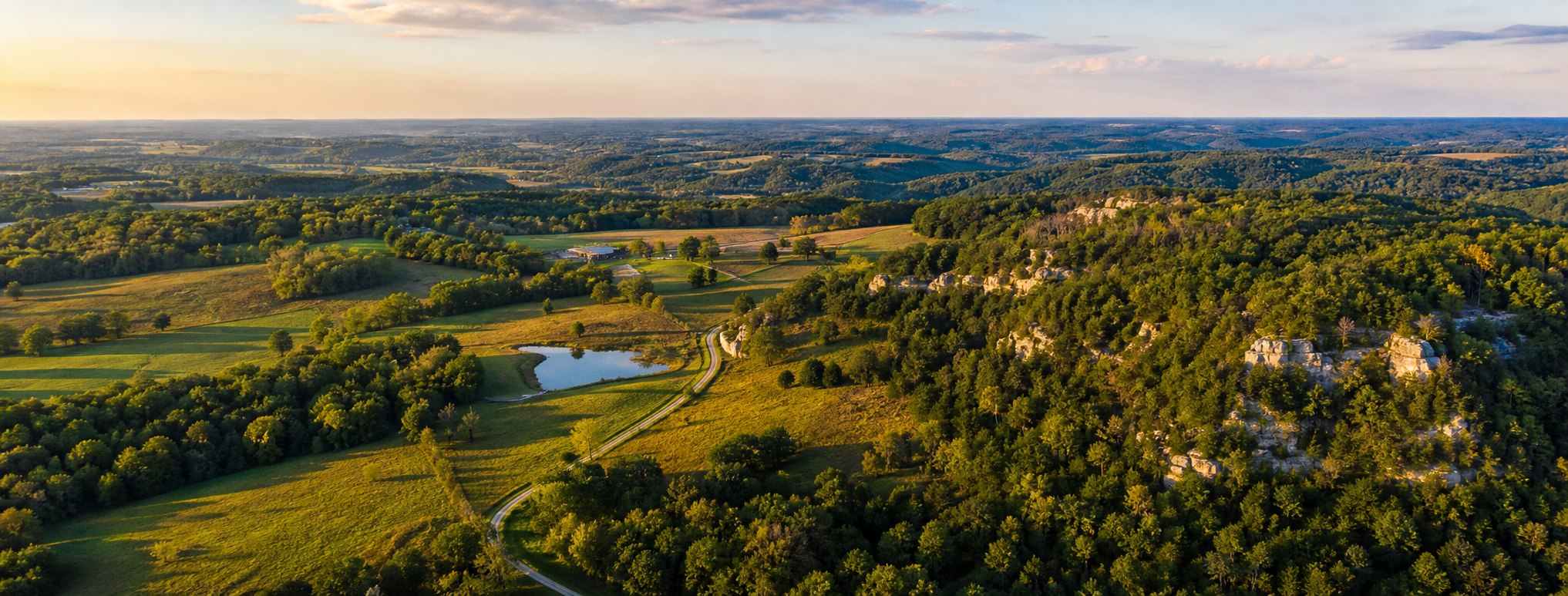 Illustrative aerial view of rolling Ozarks hills, pasture, water, and limestone outcrops representing Southwest Missouri.