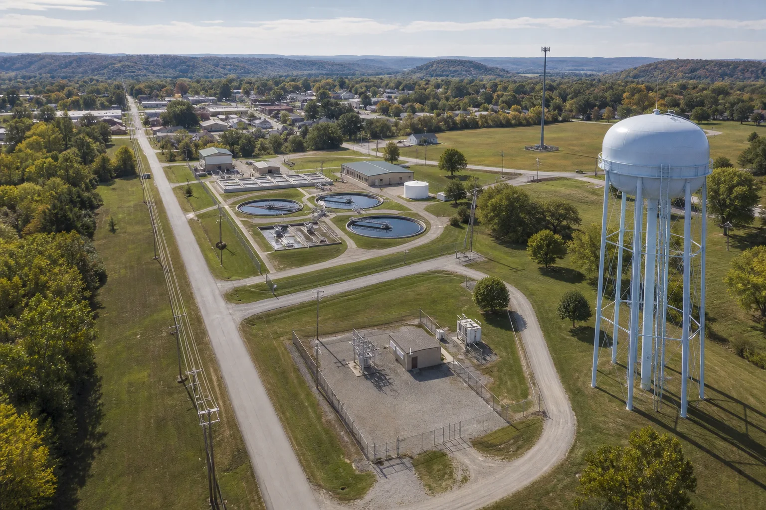 Illustrative aerial infrastructure scene showing a water tower, water treatment facility, utility pole line, electrical facility, communications tower, access routes, and surrounding site context.