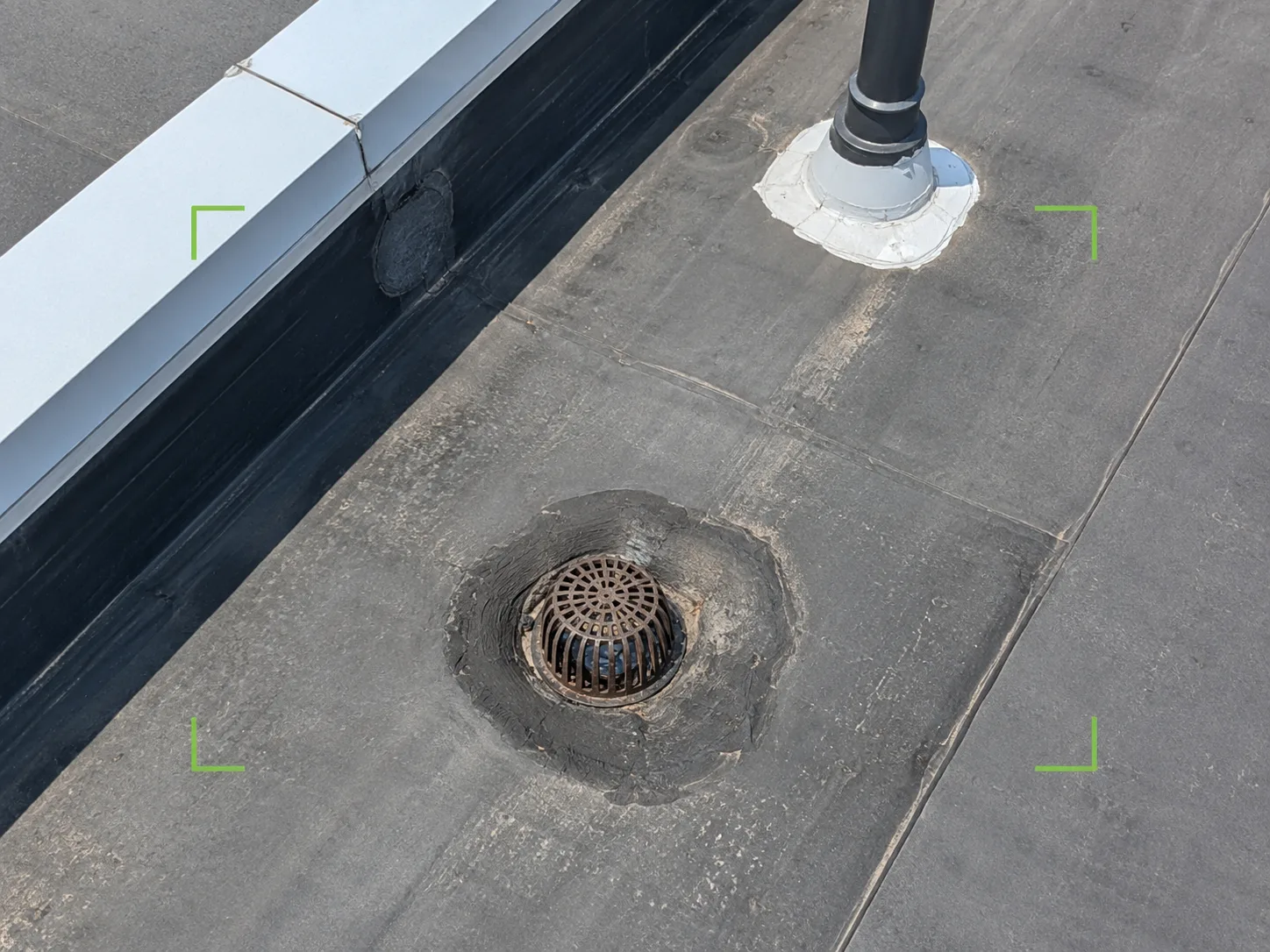 Illustrative close aerial reference of a commercial flat-roof drain, nearby membrane seams, parapet flashing, and a rooftop penetration for follow-up review.