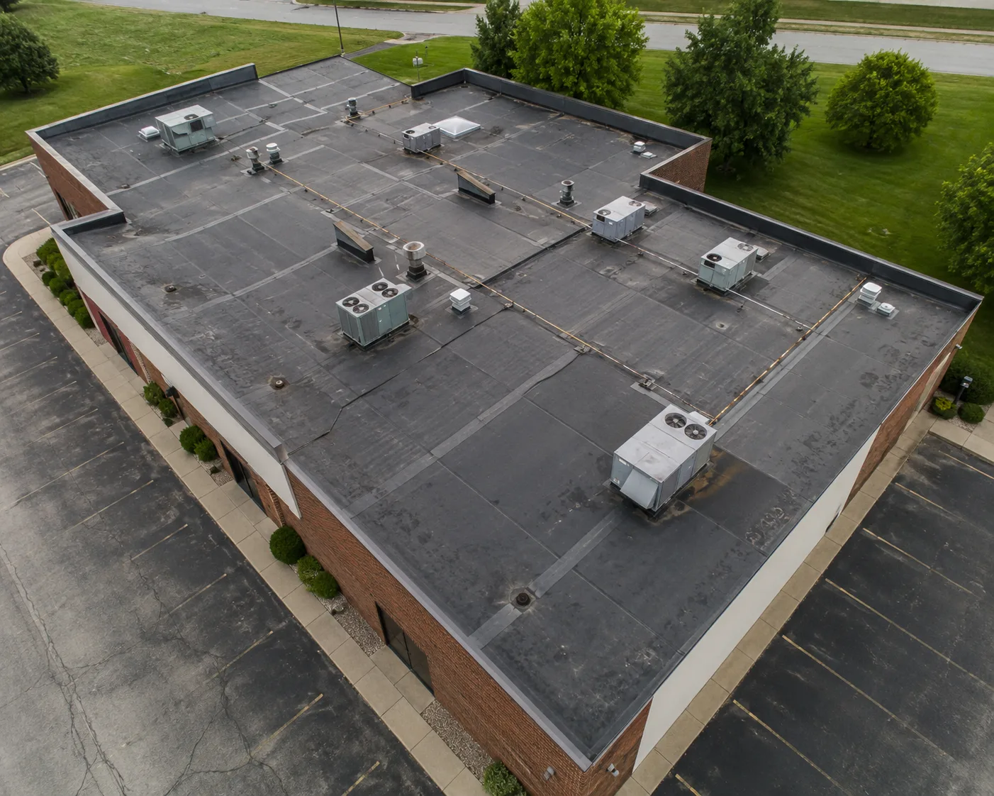 Illustrative angled aerial view of a commercial flat roof showing parapet edges, HVAC units, vents, drains, seams, and surrounding property context.